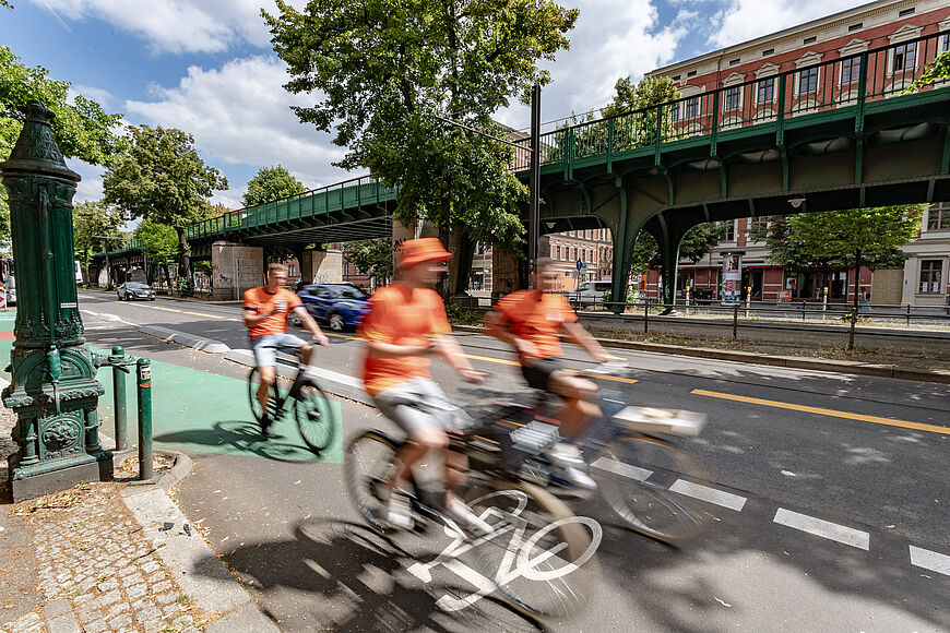 Drei Radfahrer in orangefarbenen Oberteilen fahren auf einem geschützten Radfahrstreifen.