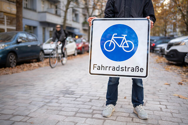 Person steht auf Straße mit Fahrradstraßen-Verkehrszeichen in den Händen, im Hintergrund fährt ein Radfahrer vorbei