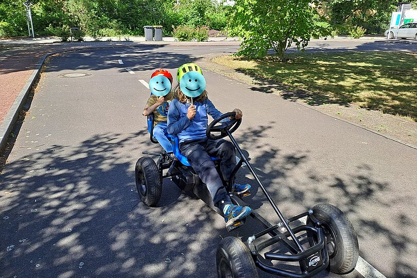 zwei Kinder sitzen zusammen auf einem Gokart. Mit lächelnden Smileys aus Papier verstecken sie ihr Gesicht.