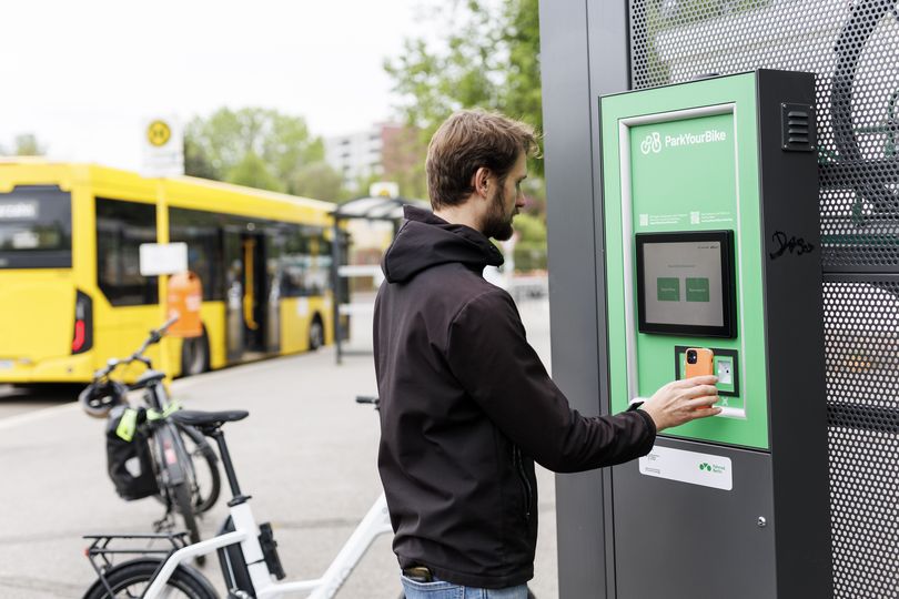 Ein Mann bedient ein Terminal an einer Sammelschließanlage für Fahrräder. Im Hintergrund steht ein Bus an einer Haltestelle.