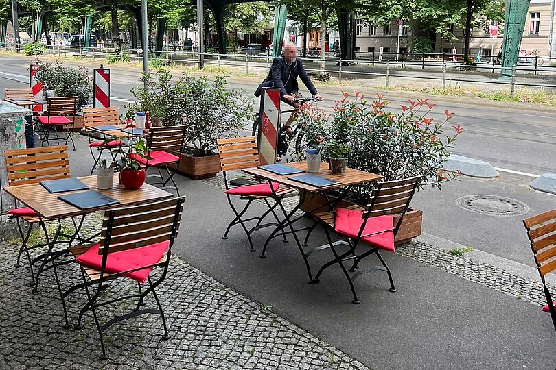 Tische und Stühle auf dem ehemaligen Radfahrstreifen, im Hintergrund fährt ein Radfahrer auf dem geschützten Radfahrstreifen