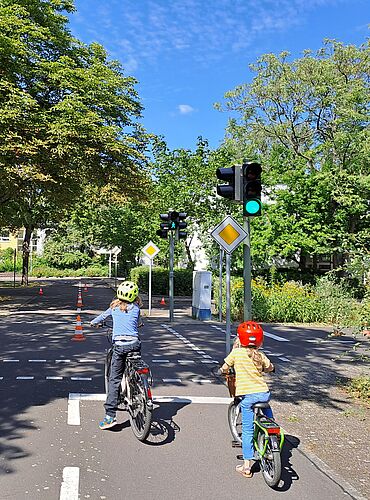 zwei Kinder sind von hinten zu sehen, wie sie auf einer Straße einer Jugendverkehrsschule mit Fahrrädern fahren