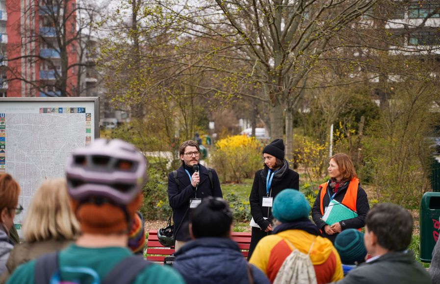 Drei Personen stehen vor einer Menge an Menschen, der Mann spricht in ein Mikrofon.