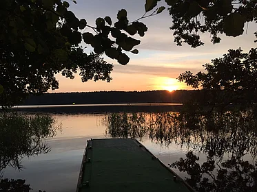 Blick auf einen See mit einem Steg, am Horizont ein Sonnenuntergang