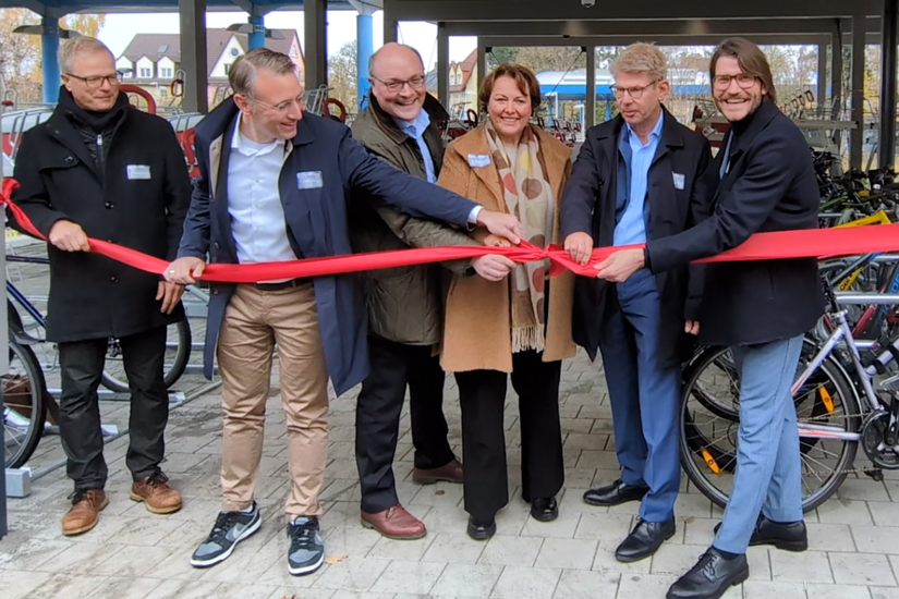 Eine Gruppe von sechs Personen öffnet feierlich ein rotes Band zur Eröffnung einer Abstellanlage für Fahrräder.