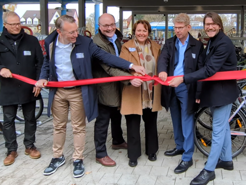 Eine Gruppe von sechs Personen öffnet feierlich ein rotes Band zur Eröffnung einer Abstellanlage für Fahrräder.