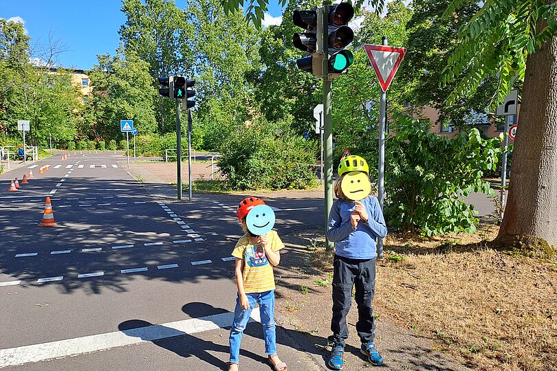zwei Kinder stehen in einer Jugendverkehrsschule am Straßenrand. Ampeln, Verkehrsschilder und Baustellenhütchen sind im Hintergrund zu sehen. Die beiden Kinder halten SMileys aus Papier vor ihr Gesicht. Das jüngere Kind hat einen lächelnden Smiley, das ältere Kind hat einen neutral schauenden Smiley
