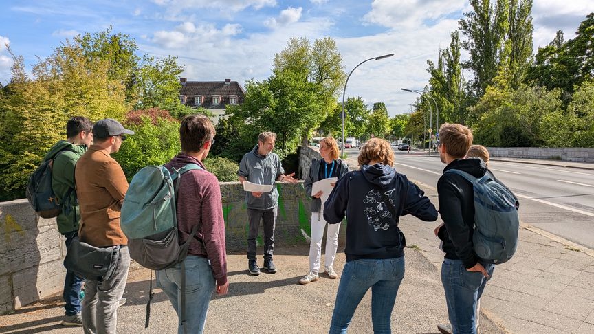 mehrere Personen stehen an einer Straße im Halbkreis und hören einem Vortrag zu