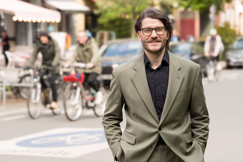 Mann blickt in die Kamera. Im Hintergrund ist eine Fahrradstraße mit zwei Radfahrenden zu sehen.