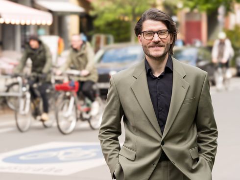 Mann blickt in die Kamera. Im Hintergrund ist eine Fahrradstraße mit zwei Radfahrenden zu sehen.