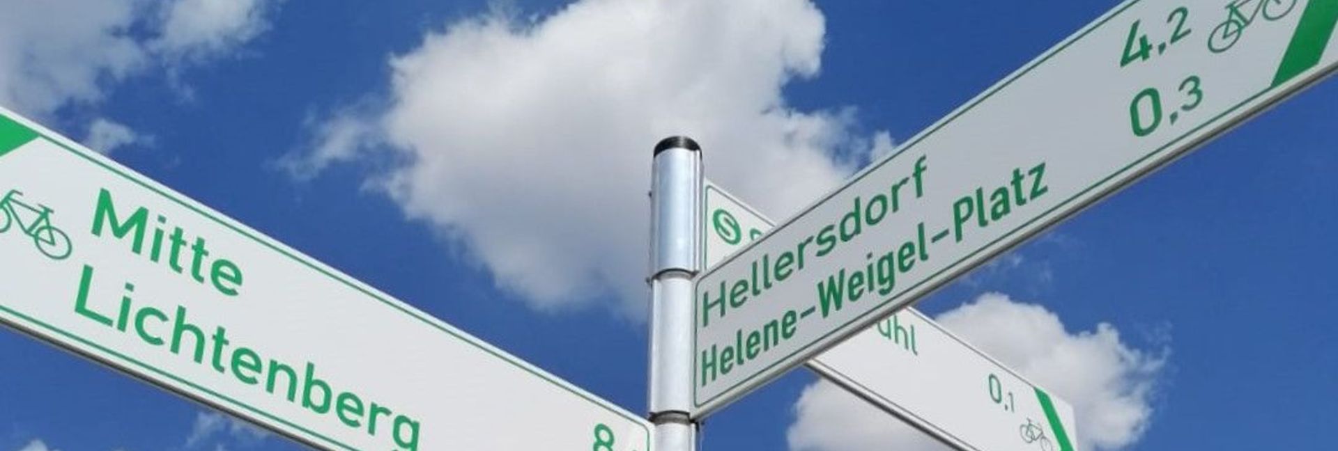 Radwegweiser zeigen nach Mitte und Lichtenberg sowie nach Hellersdorf. Im Hintergrund blauer Himmel und Wolken.