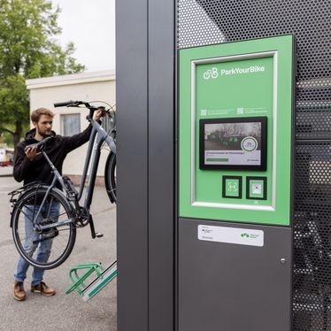 Ein Mann parkt sein Fahrrad in einer Doppelstockanlage, im Vordergrund ist ein grünes Terminal von ParkYourBike zu sehen.