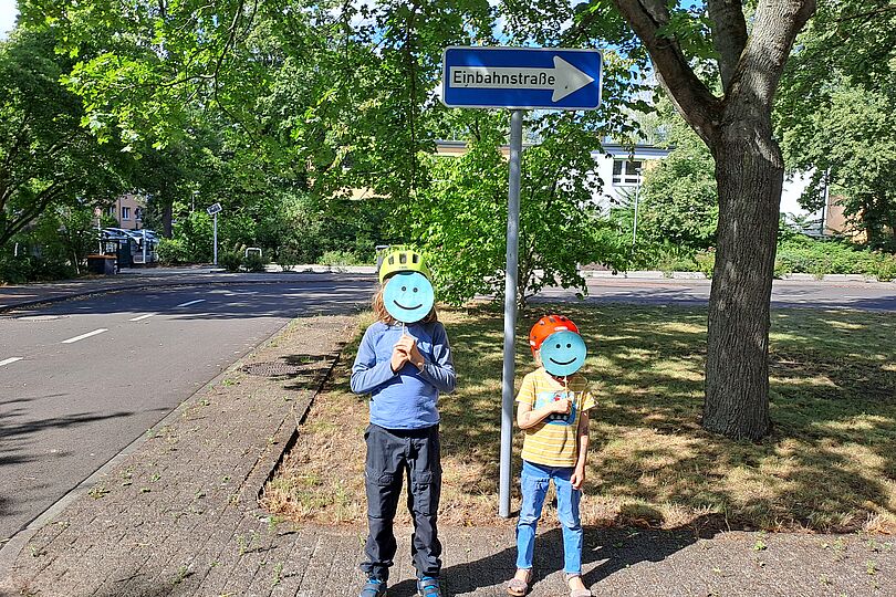 zwei Kinder mit Fahrradhelmen stehen in einer Jugendverkehrsschule vor einem Einbahnstraßen-Schild und halten lächelnde Smileys aus Papier vor ihr Gesicht.