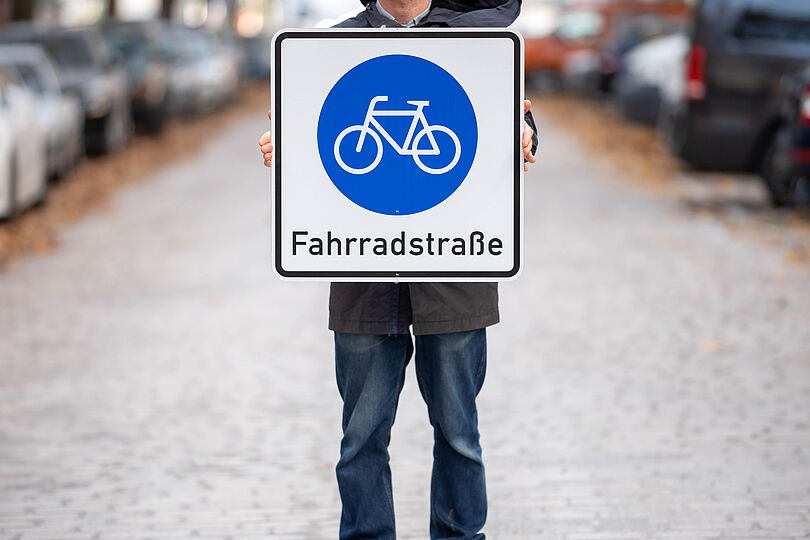Eine Person steht auf der Straße und hält das Verkehrsschild einer Fahrradstraße in beiden Händen.