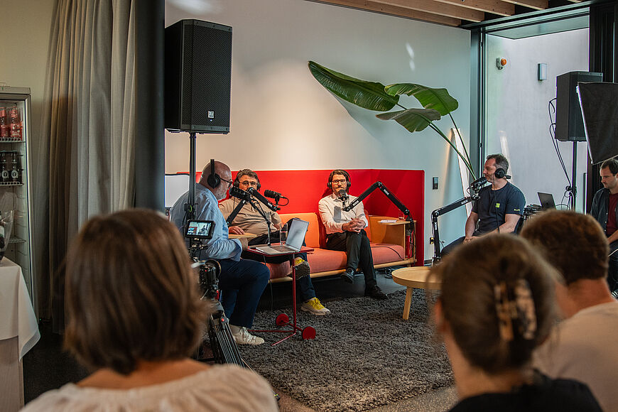 Live-Aufzeichnung eines Podcasts, Publikum schaut zu vier Personen mit Kopfhörern und Mikrofonen, die einen Podcast aufzeichnen