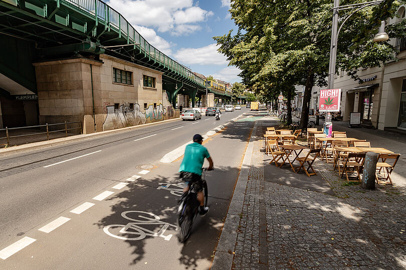 Fahrradfahrer fährt auf einem geschützten Radfahrstreifen. Auf dem Gehweg rechts von ihm stehen Tische und Stühle eines Cafés.