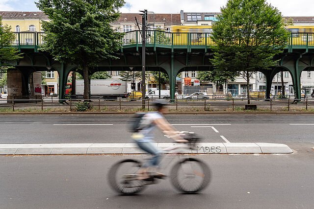 Fahrradfahrer fährt auf einem geschützten Radweg, im Hintergrund ist das grüne Viadukt der Schönhauser Allee zu sehen, auf dem eine gelbe U-Bahn fährt.