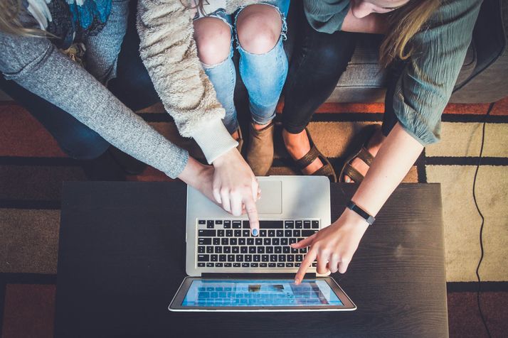 drei Menschen sitzen vor einem Laptop, man sieht nur ihre Arme und Hände, die alle auf den Bildschirm zeigen