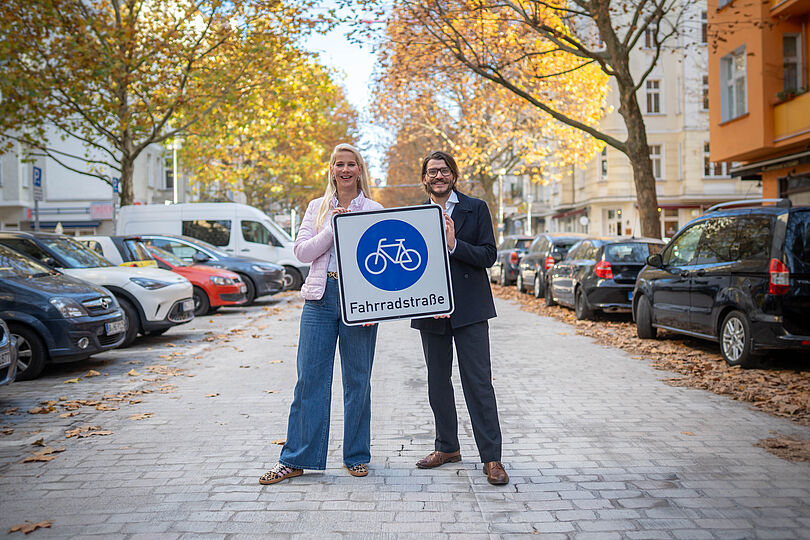 Manuela Anders-Granitzki und Michael Fugel stehen auf der Hufelandstraße und halten einen Fahrradstraßen-Verkehrsschild in den Händen