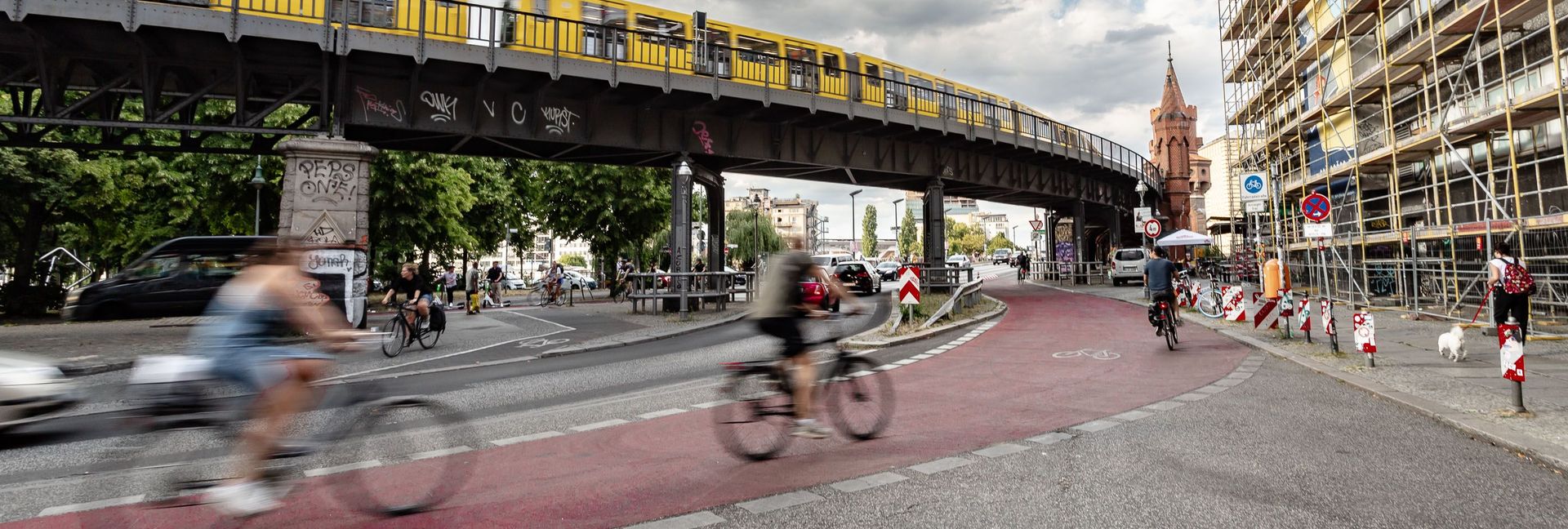 Zu sehen sind Fahrradfahrer auf einem rotmarkierten Radfahrstreifen. Im Hintergrund ist eine Brücke mit gelber Bahn zu sehen.