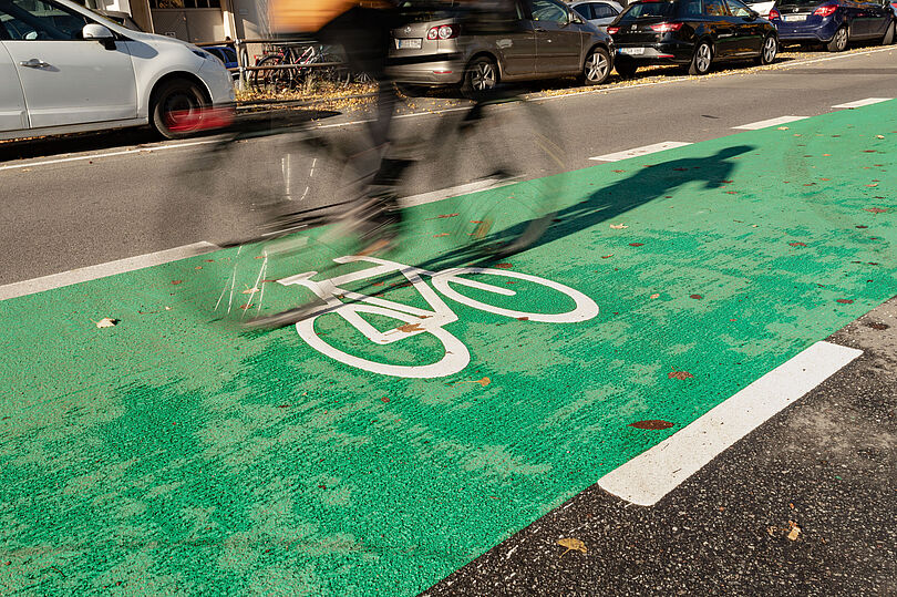 Detailaufnahme eines grün markierten Schutzstreifens mit weißem Fahrradpiktogramm. Auf dem Schutzstreifen fährt ein Fahrrad.