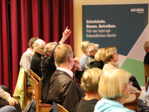 Blick von der Seite auf Publikum in Stuhlreihen, eine Person meldet sich