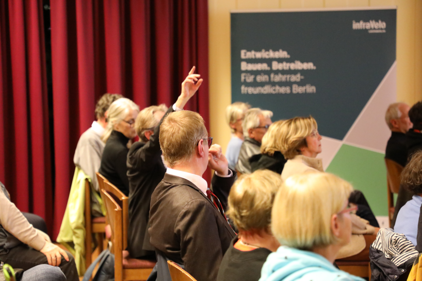 Blick von der Seite auf Publikum in Stuhlreihen, eine Person hat die Hand gehoben