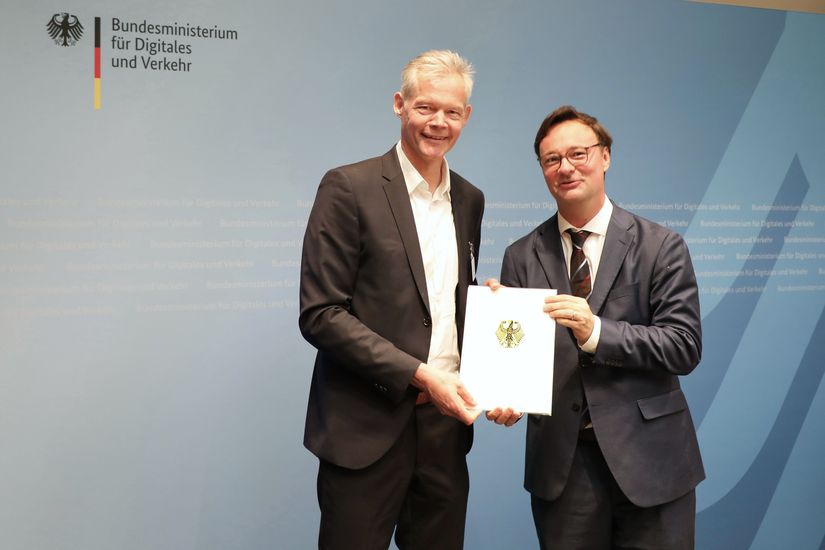 Zwei Männer halten Dokument in die Kamera vor Fotowand "Bundesministerium für Digitales und Verkehr"