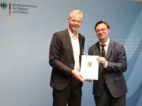 Zwei Männer halten Dokument in die Kamera vor Fotowand "Bundesministerium für Digitales und Verkehr"