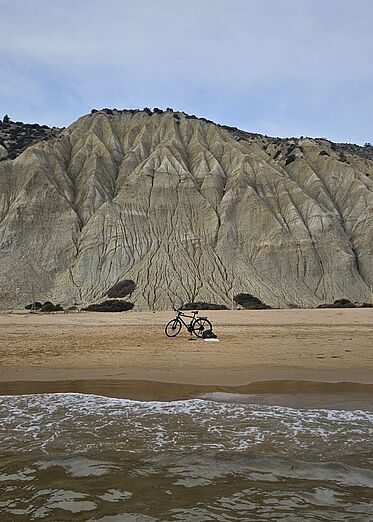 Fahrrad steht am Strand, im Vordergrund das Meer, im Hintergrund Berge