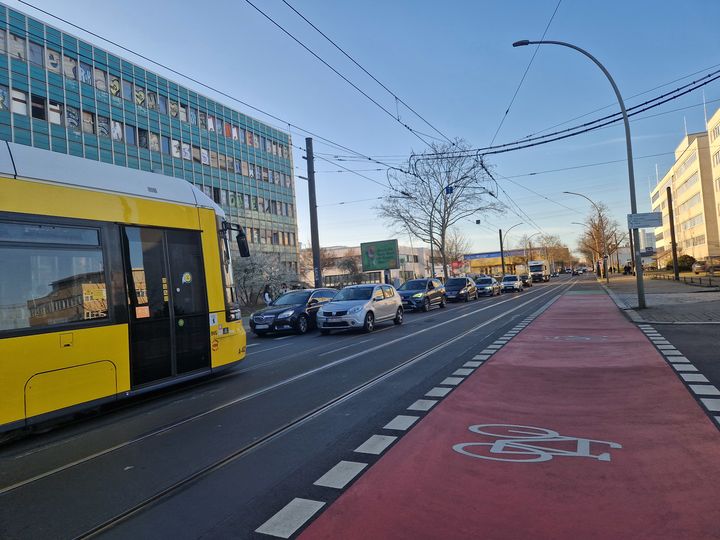 Rot beschichteter Schutzstreifen entlang der Straßenbahngleise mit einem Fahrradpiktogramm.