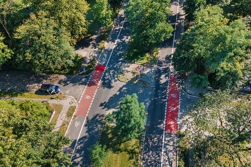 Blick von oben auf eine Straße mit einem teilweise rot markierten Radfahrstreifen auf beiden Seiten, in der Mitte ein begrünter Mittelstreifen, viele grüne Bäume