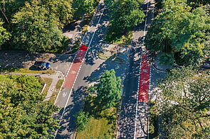 Blick von oben auf eine Straße mit einem teilweise rot markierten Radfahrstreifen auf beiden Seiten, in der Mitte ein begrünter Mittelstreifen, viele grüne Bäume
