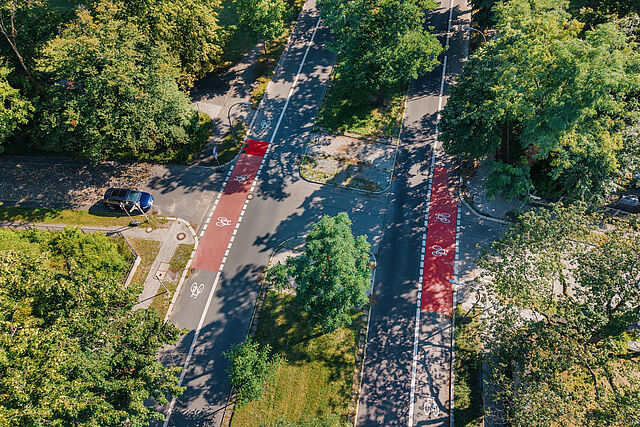 Blick von oben auf eine Straße mit einem teilweise rot markierten Radfahrstreifen auf beiden Seiten, in der Mitte ein begrünter Mittelstreifen, viele grüne Bäume