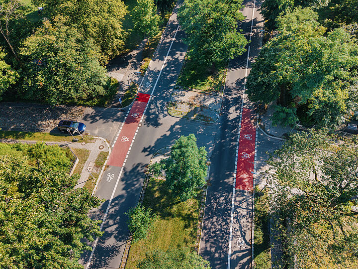 Blick von oben auf eine Straße mit einem teilweise rot markierten Radfahrstreifen auf beiden Seiten, in der Mitte ein begrünter Mittelstreifen, viele grüne Bäume