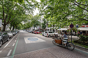 Radfahrerin mit einem Lastenrad auf einer markierten Fahrradstraße. Auf der rechten Fahrbahnrand sind Kreuzberger Bügel als Fahrradabstellanlagen angebracht.