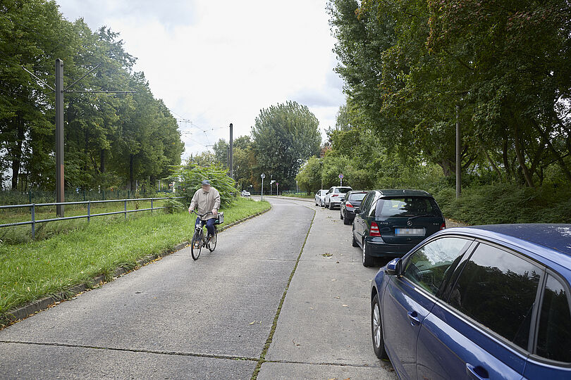 Radfahrende auf einer Straße ohne Radverkehrsinfrastruktur und mit parkenden Autos auf der rechten Straßenrand.