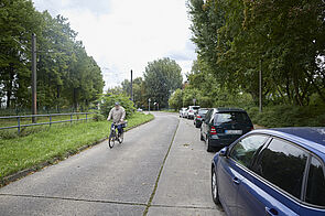 Radfahrende auf einer Straße ohne Radverkehrsinfrastruktur und mit parkenden Autos auf der rechten Straßenrand.