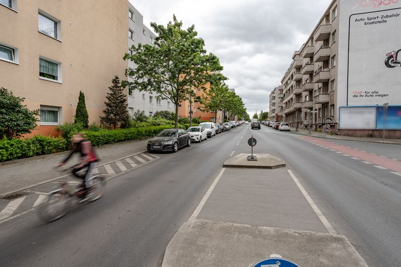 Radfahrende auf einer Autostraße ohne Radweg mit parkenden Autos und einem Fußgängerüberweg.