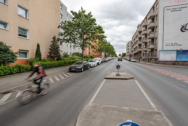 Radfahrende auf einer Autostraße ohne Radweg mit parkenden Autos und einem Fußgängerüberweg.
