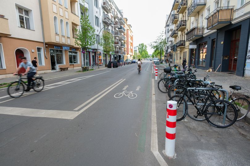 Radfahrende auf einer markierten Fahrradstraße mit rotweißen Pollern, Fahrradpiktogrammen und Kreuzbergerbügeln als Fahrradabstellplätze.