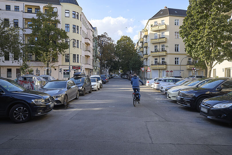 Radfahrende auf einer Straße ohne Radverkehrsinfrastruktur und mit parkenden Autos auf beiden Straßenseiten.