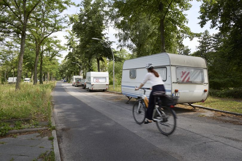 Radfahrerin auf baumbestandener Berliner Straße mit parkenden Wohnmobil-Anhängern und ohne gesonderte Radverkehrsinfrastruktur – eingeschränkte Verkehrssicherheit und mangelnde Radwege im grünen Stadtbereich.