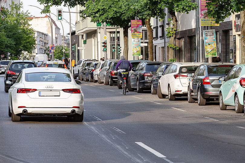 Autostraße ohne einen Fahrradweg mit Autoverkehr und auf der rechten Seite mit parkenden Autos und einem Radfahrenden.
