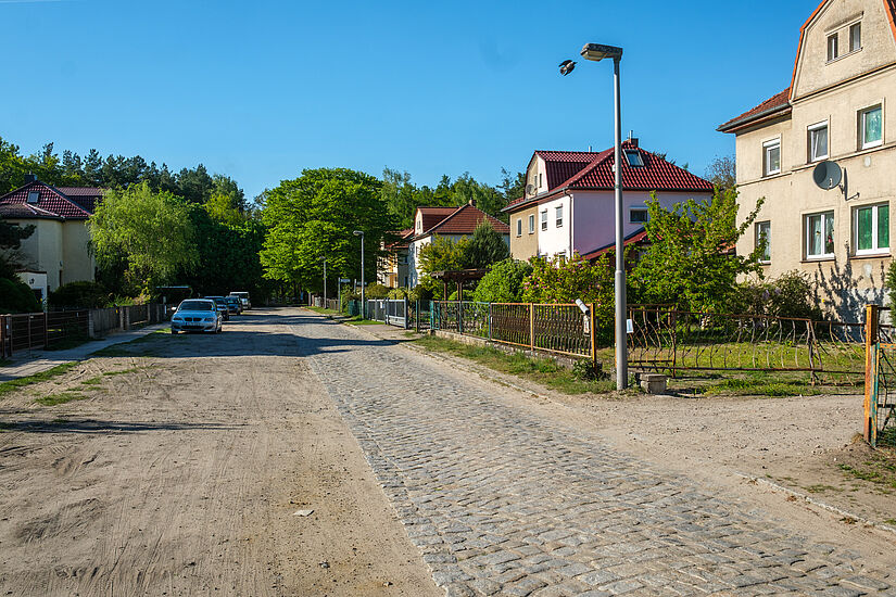 Kopfsteinpflasterstraße neben einem Schotterweg durch ein Wohngebiet mit Einfamilienhäusern.
