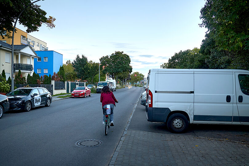 Radfahrerin auf einer Autostraße ohne Radverkehrsinfrastruktur. Auf beiden Seiten der Straße sind Autoparkplätze.