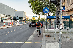 Radfahrende auf einem schmalen Hochboardradweg, der einem U-Bahneingang vorbeiführt.