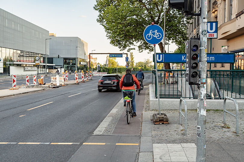 Radfahrende auf einem schmalen Hochboardradweg, der einem U-Bahneingang vorbeiführt.