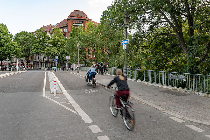 Radfahrende auf der durch Betonborde geschütztem Radweg auf der Hobrechtbrücke.