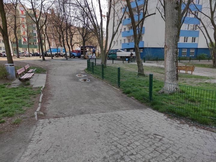 Teilweise unbefestigter Weg entlang eines Spielplatzzaunes.